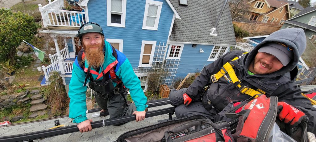 Two solar installers are smiling at the camera while working on the roof of a home